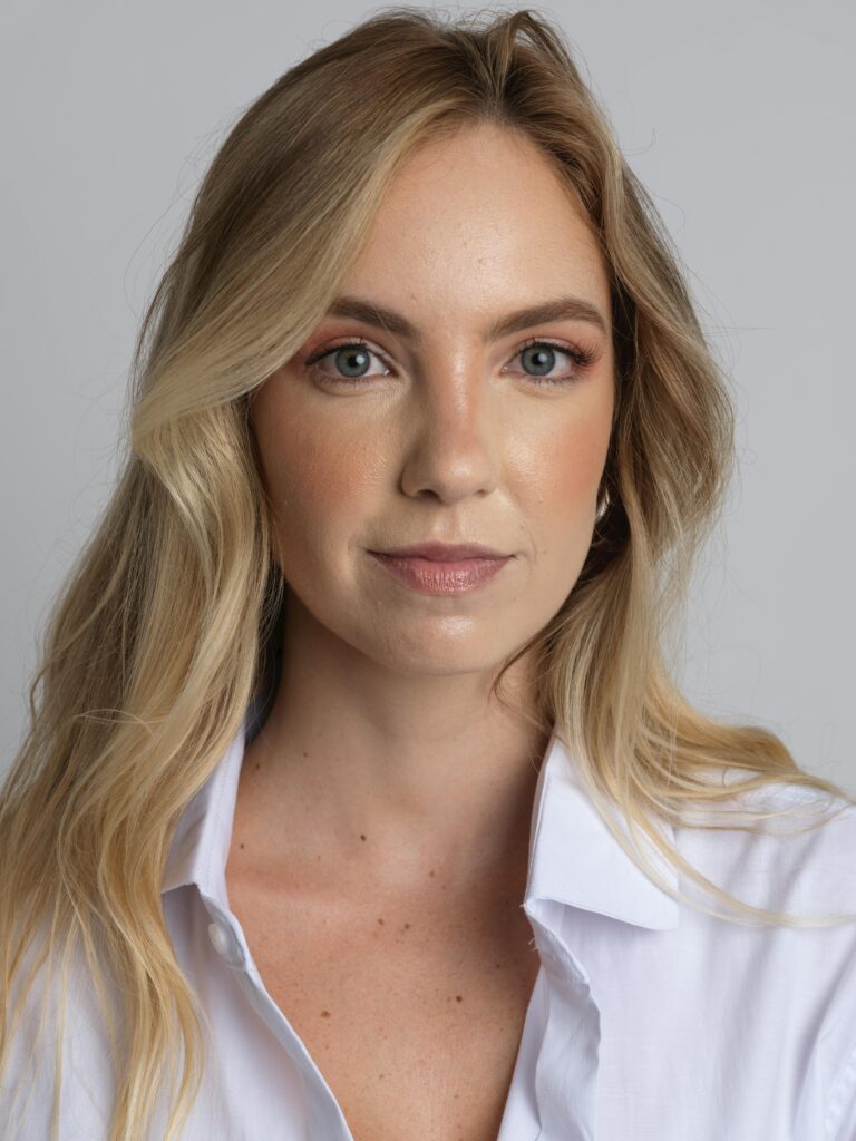A confident blonde woman posed against a neutral background wearing a white shirt.