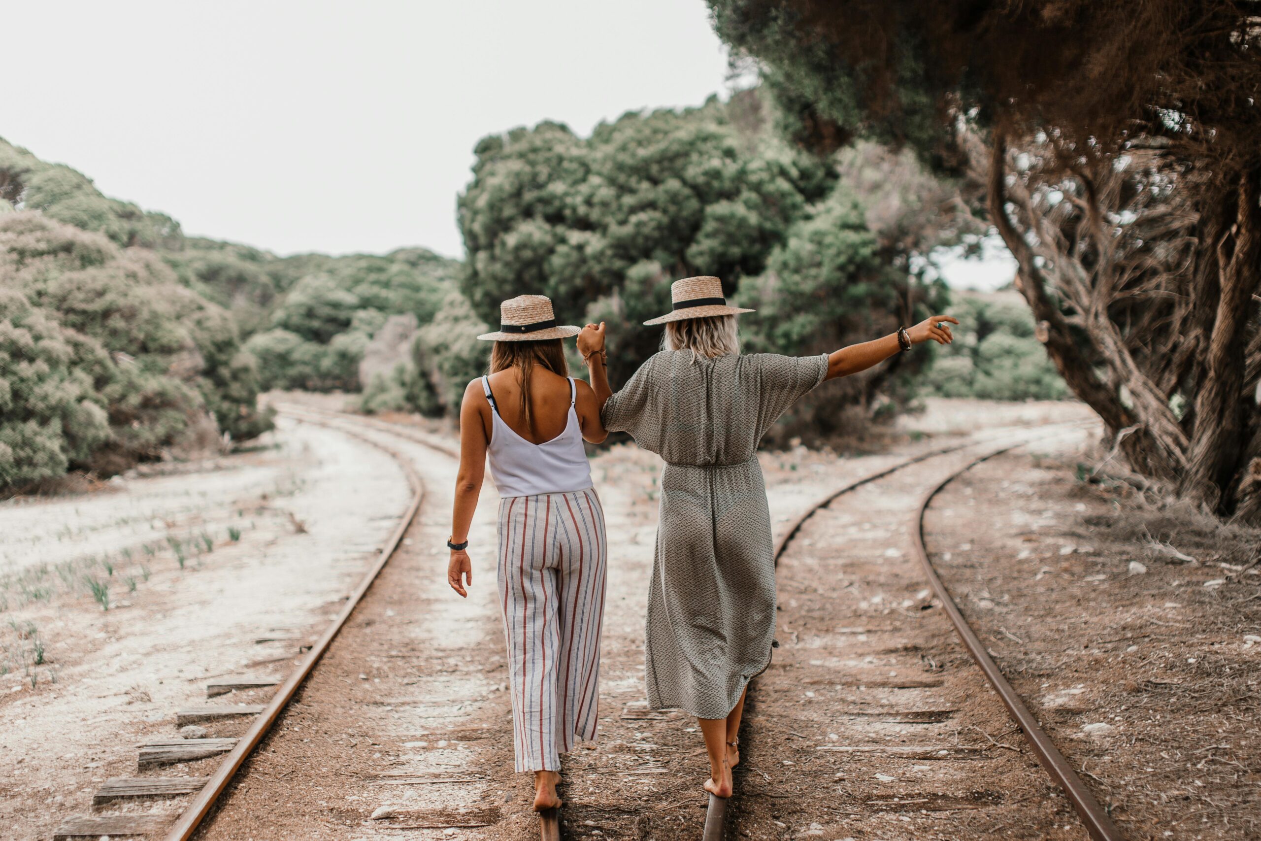 Two women holding hands as they walk barefoot on railway tracks in a countryside setting.
