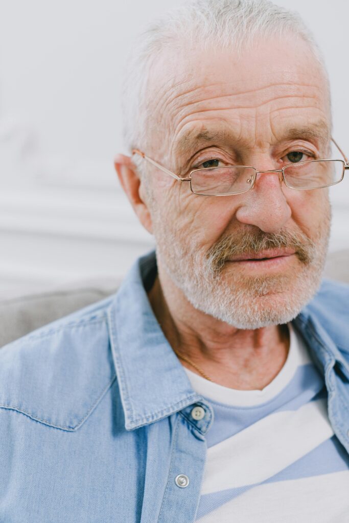 Elderly man with gray hair and eyeglasses sitting indoors, wearing casual denim shirt.